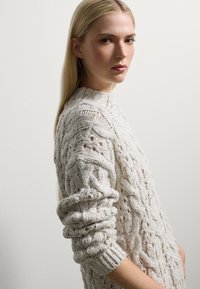 Pull en maille de couleur blanc cassé avec un motif de câble texturé, col montant et poignets roulés. La laine épaisse ajoute de la profondeur et un intérêt visuel.