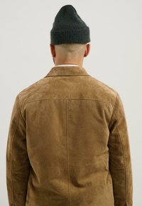Mann, der eine braune Wildlederjacke und eine dunkle Strickmütze trägt, von hinten vor einem schlichten hellen Hintergrund gesehen.
