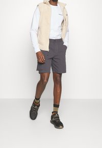 Beige fleece-vest, vit vit långärmad skjorta, mörkgrå cargoshorts och svarta skor med grå detaljer och bruna strumpor. Slät textur, bekväm passform.