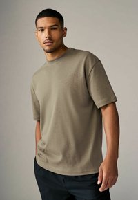 Camiseta de algodón verde olivo con mangas cortas, cuello redondo, corte relajado y textura suave. Combinada con pantalones negros.