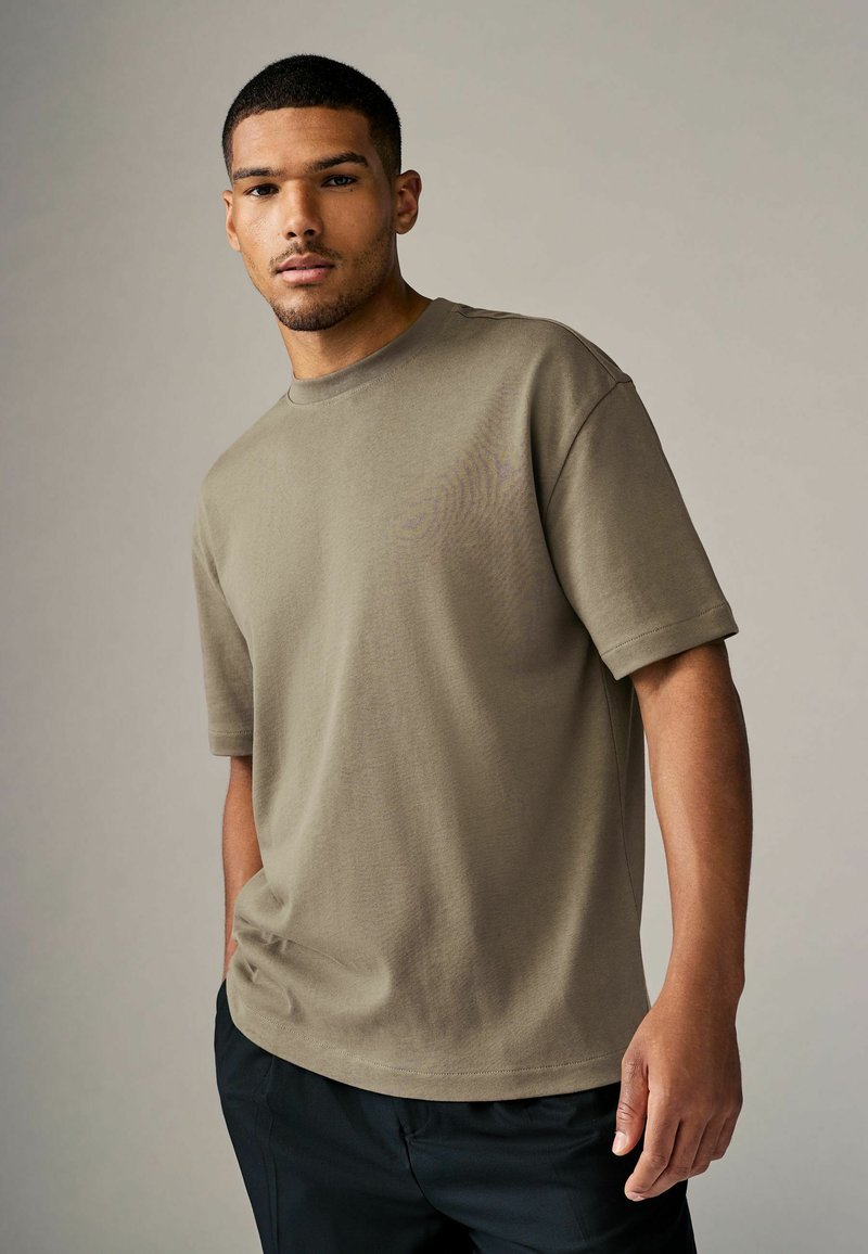 Camiseta de algodón verde olivo con mangas cortas, cuello redondo, corte relajado y textura suave. Combinada con pantalones negros.