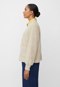 Beżowa polarowa kurtka o prostym kroju, z kieszeniami bocznymi i ściągaczami, noszona na żółtej koszuli z kołnierzykiem. Gładka tekstura i casualowy design.