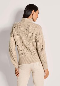 Frau mit langen, dunklen Haaren, die einen beigen, grob gestrickten Rollkragenpullover und hellbeige Hosen mit einer gestickten "M" auf der Rückentasche trägt und von der Kamera wegschaut.
