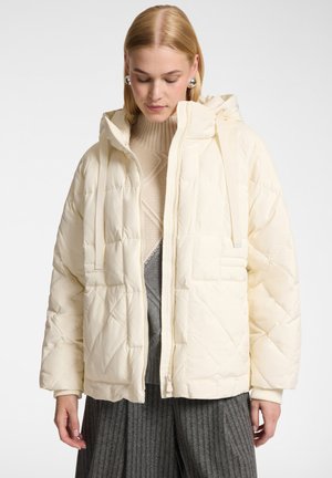 Donna con capelli biondi corti che indossa grandi orecchini d'argento, giacca trapuntata crema con cappuccio, maglione beige e pantaloni grigi a coste, guarda verso il basso.