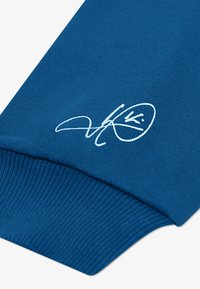 Modra majica s kapuco z ribanim robom in manšetami, izdelana iz mehkega blaga, s beli podpisom natisnjenim na rokavu.