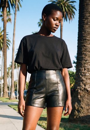 Mujer de cabello corto con una camiseta negra y shorts de cuero negro de talle alto, de pie al aire libre cerca de palmeras en un día soleado.