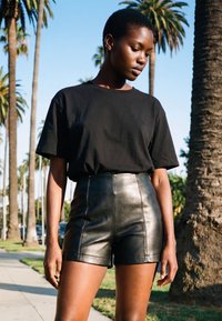 Sieviete ar īsiem matiem, valkājot melnu T-kreklu un augsta vidukļa melnas ādas šortus, stāv ārā pie palmu kokiem saulainā dienā.