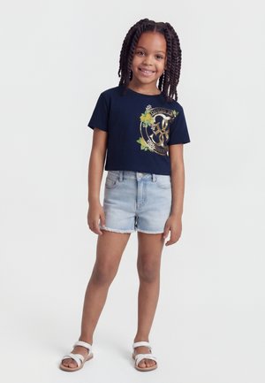 Usmievavá dievčina s vrkočmi, ktorá má na sebe námornícky modré tričko s kvetinovým logom, svetlé rifľové šortky a biele sandále, stojaca pred jednoduchým pozadím.