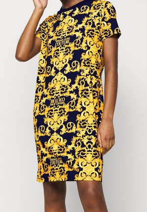 Granatowa sukienka z krótkim rękawem z jaskrawożółtymi ozdobnymi wzorami barokowymi oraz napisem „Versace Jeans Couture”, noszona przez osobę.