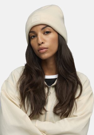 Cream ribbed beanie with a square logo patch, paired with a light beige zip-up jacket and a black and white layered shirt beneath.