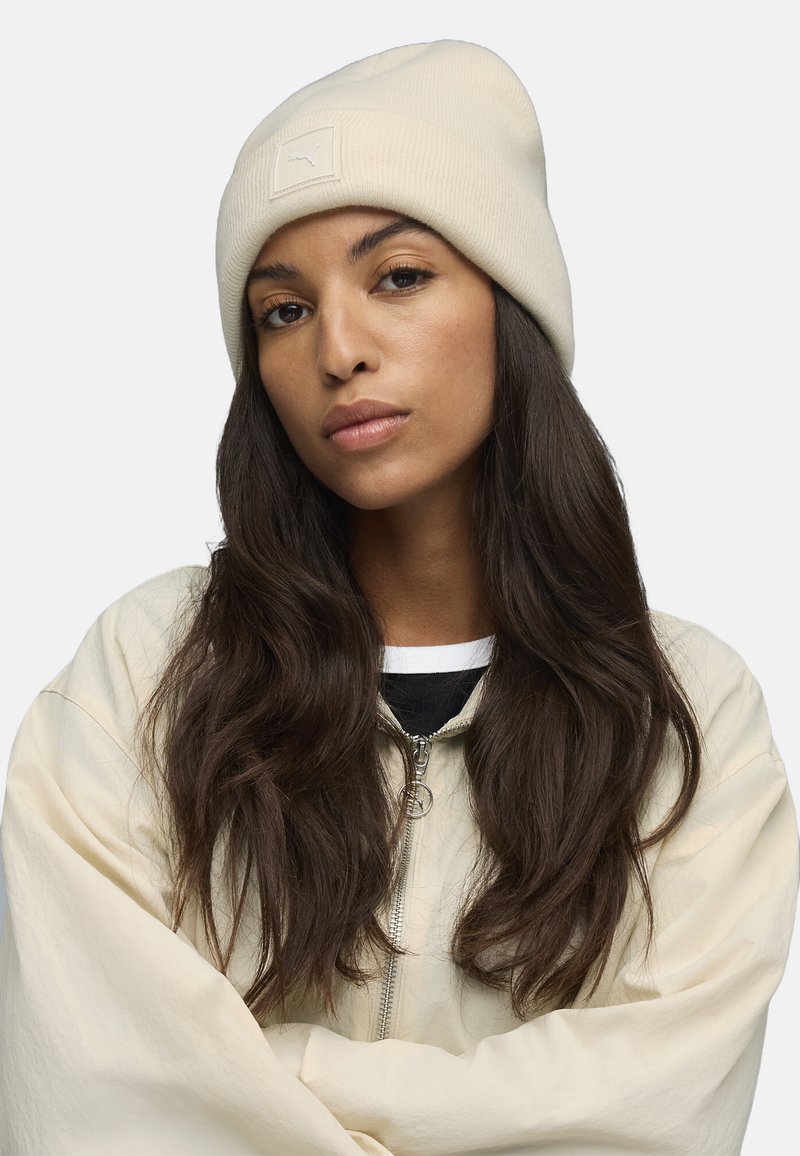 Gorro de canalé crema con un parche de logo rectangular, combinado con una chaqueta de cremallera color beige claro y una camiseta de capas en negro y blanco debajo.