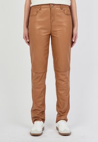 Bruine leren broeken met een gladde textuur, met een slim fit ontwerp, voorzakken en gedetailleerde stiksels op de knieën. Gepaard met witte sneakers.