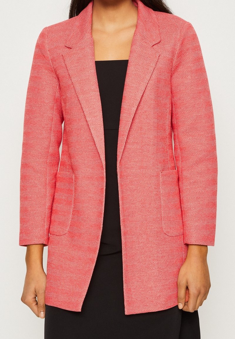 Femme portant un blazer rose à motif chevrons sur une robe noire, montrée du cou jusqu'au milieu de la cuisse, les bras détendus le long du corps.