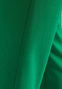 Detailní záběr na hladkou, živou zelenou látku s viditelným švem a jemnými záhyby, který zdůrazňuje texturu a kvalitu materiálu.