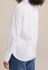 Vit långärmad skjorta i mjuk bomull, med en böjd nederkant och knäppte manschetter, tillsammans med blå denimjeans.