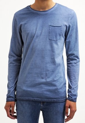 Homme portant une chemise bleu ajustée à manches longues avec une poche poitrine et un jean bleu, debout devant un fond blanc uni.