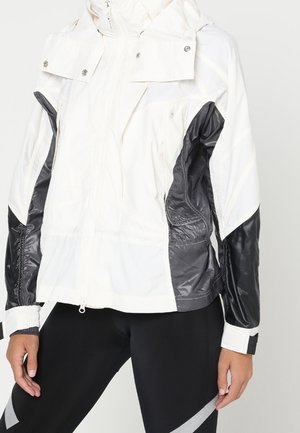Veste coupe-vent à capuche blanche et noire portée avec des leggings noirs présentant des bandes diagonales réfléchissantes.