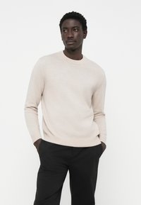 Suéter beige de cuello redondo hecho de tela suave, con mangas largas y puños acanalados, combinado con pantalones oscuros. Sin patrones ni acentos.