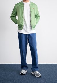 Giacca bomber verde chiaro con zip, t-shirt bianca a girocollo, jeans in denim blu e sneakers grigie. Design semplice, vestibilità casual.