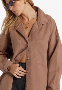 Bruine oversized button-up blouse gemaakt van lichtgewicht stof, met twee borstzakken en subtiele textuur. Zwarte broek zichtbaar bij de taille.