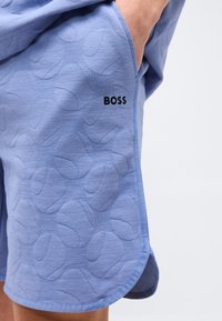 Ανοιχτό μπλε σορτς με ανάγλυφα σχέδια τεννιστικών μπαλών και λογότυπο "BOSS", κοντινή φωτογραφία χεριού στην πλαϊνή τσέπη σε απλό φόντο.