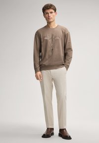 Felpa marrone con un design floreale testurizzato, abbinata a pantaloni beige chiaro e stivali marroni scuri, che mostrano una vestibilità rilassata e una palette di colori neutri.