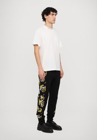 Balta T-krekls ar īsām piedurknēm un kabatu, apvienots ar melniem sporta biksēm, kuru sānos ir dzelteni un baltā krāsā ornamenti. Melni apavi.