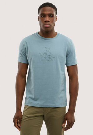 Uomo che indossa una maglietta Trussardi azzurro chiaro con logo ricamato e pantaloni verde oliva, in piedi davanti a uno sfondo neutro.