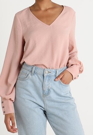 Kobieta ubrana w jasnoróżową bluzkę z długim rękawem i mankietami na guziki oraz jasnoniebieskie dżinsy z wysokim stanem, ręka w kieszeni.