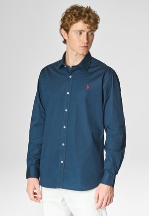 Giovane uomo con capelli ricci che indossa una camicia blu navy con un logo rosso, abbinata a pantaloni bianchi, che guarda di lato.