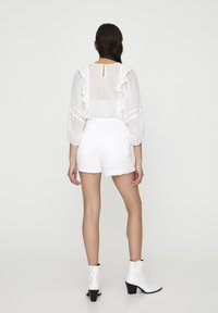Femme debout de dos portant une blouse blanche à manches longues avec des volants, un short blanc taille haute et des bottines blanches à talons blocs.