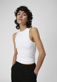 Débardeur blanc côtelé au design ajusté, sans manches et avec une longueur crop, associé à un pantalon noir. Esthétique simple et épurée.