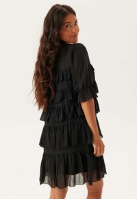 Svart lagd klänning med volanglager, semi-genomskinligt tyg, korta puffärmar och en figursydd överdel, vilket framhäver en flödande silhuett.