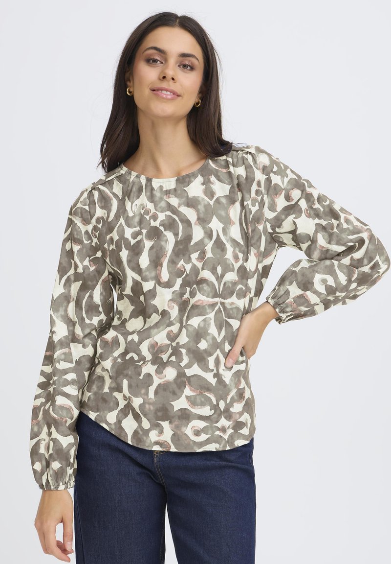 Femme portant une blouse à manches longues avec un motif abstrait beige et gris, associée à un jean bleu foncé, posant avec une main sur la hanche.