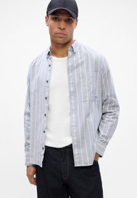 Pelēka un balta svītraina pogājama krekls, garām piedurknēm, ar vienu krūšu kabatu, valkāts vaļīgi virs baltā T-kreklu, kopā ar tumšiem džinsiem.