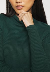 Pull à col roulé côtelé en tissu vert foncé, avec manches longues et coupe ajustée qui met en valeur la texture et le motif.