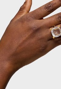 Złoty pierścionek z dużym, fasetowanym przezroczystym kamieniem, noszony na dłoni o teksturowanej skórze, ukazującej naturalne odcienie.