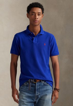 Jeune homme portant un polo bleu avec un logo rouge, un jean bleu clair, une montre marron et une ceinture en tissu rayée, debout devant un fond uni.