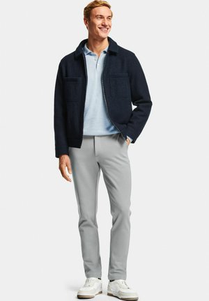 Homme souriant debout portant une veste bleue marine, un polo bleu clair, un pantalon gris clair et des baskets blanches sur un fond blanc uni.