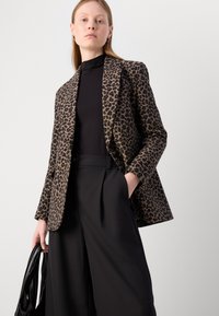 Leopardmuster-Blazer aus braunem und schwarzem Stoff, taillierte Passform mit spitzen Revers. Kombiniert mit hochgeschnittenen schwarzen Hosen und einer schwarzen Tasche.