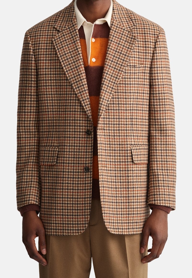 Hombre vistiendo un blazer de pata de gallo marrón y beige sobre una camisa con cuello y un suéter a rayas en marrón, naranja y crema, combinado con pantalones color coñac.