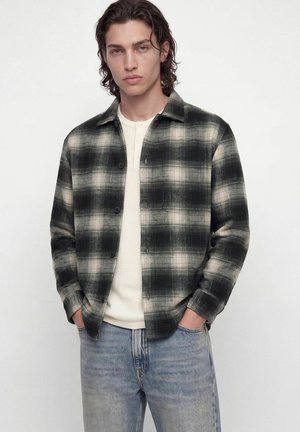 Jeune homme aux cheveux mi-longs portant une chemise à carreaux noire et crème boutonnée par-dessus un henley blanc et un jean bleu clair, les mains dans les poches.
