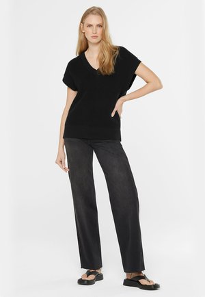 Blonde woman wearing black knit sleeveless V-neck sweater, wide-leg black jeans, and black sandals, standing with hand on hip.