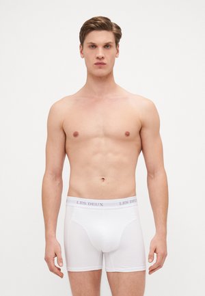 Weiße Baumwoll-Boxershorts mit weicher Textur, ausgestattet mit einem grauen Logo-Bund und einem mittellangen Schnitt. Keine Muster. Das Modell ist oberkörperfrei.