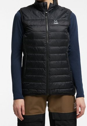 Persoon draagt een zwart doorgestikt vest over een donkerblauw hemd met lange mouwen en bruine en zwarte broek, staat met ontspannen armen naast het lichaam.