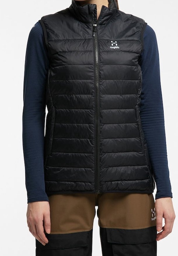 Personne portant un gilet matelassé noir par-dessus une chemise bleu marine à manches longues et un pantalon marron et noir, debout les bras détendus le long du corps.