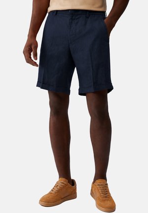 Uomo che indossa pantaloncini blu scuro con orli risvoltati e sneakers in camoscio beige, in piedi con una mano in tasca davanti a uno sfondo semplice.
