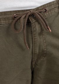 Brown drawstring tied in a bow on olive green pants worn with a white shirt visible above the waistband.