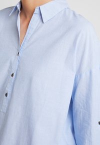 Camicia button-up azzurro chiaro con colletto, caratterizzata da un tessuto testurizzato, una leggera lucentezza e bottoni marroni lungo la chiusura.