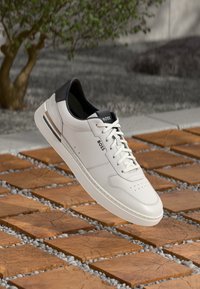 White leather sneaker with black heel accent, lace-up design, perforated toe, and branding. Elevated above a patterned stone surface.